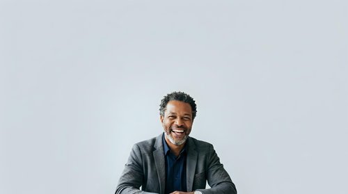 Middle-aged man with dark skin and short dark hair, wearing a charcoal blazer over a navy shirt, smiling warmly at camera against neutral background for corporate and professional use.