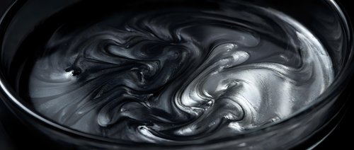 Dark swirling liquid paint creates mesmerizing abstract patterns in black bowl. Fluid art texture with metallic sheen for creative design projects.