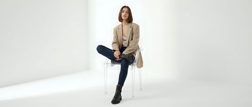 Young professional woman in beige blazer and dark jeans sitting on white chair against neutral background for business and lifestyle concepts.