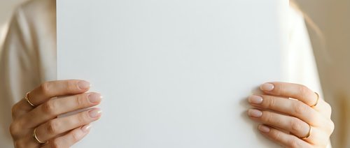 Hands of a middle-aged woman with medium skin tone holding blank white paper or document, wearing gold rings, against neutral beige background for mockup and presentation purposes.