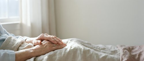 Caring hands of elderly couple holding together in bed showing love, support and companionship in comfortable bedroom setting with soft natural lighting.