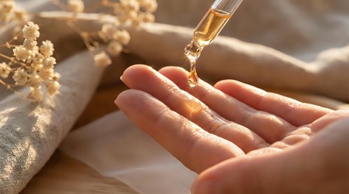 Golden serum drops from pipette into cupped hands with dried flowers on neutral fabric background for skincare and beauty wellness routine.