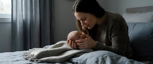 Young mother tenderly holding and kissing newborn baby while sitting on bed in cozy bedroom. Intimate bonding moment between parent and infant in soft natural light.