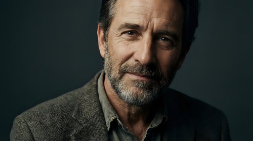 Mature Caucasian businessman with gray beard smiling confidently in professional portrait against dark background for corporate marketing materials.