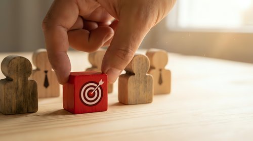 Hand placing a red wooden block with target and arrow icon among human figure blocks, symbolizing leadership, recruitment, and business strategy.