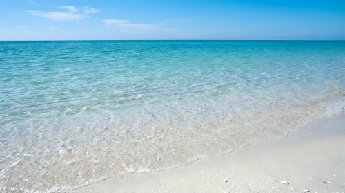 Pristine tropical beach with crystal clear turquoise water gently lapping white sand shore under bright blue sky with soft clouds for vacation travel.
