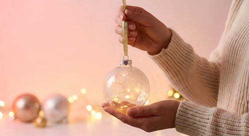 Female hands in cozy knit sweater holding glowing Christmas ornament with warm fairy lights and festive decorations on soft pink background for holiday design.