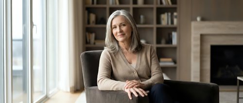 Confident mature businesswoman with gray hair sitting at desk in modern office interior with bookshelf and fireplace, professional corporate executive portrait.