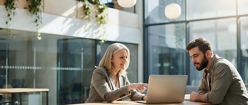 Professional blonde businesswoman and bearded male colleague collaborating on laptop in modern office with natural lighting and glass architecture.