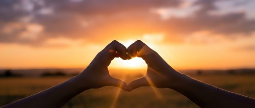 Hands forming heart shape silhouette against golden sunset sky with warm sunlight streaming through fingers creating romantic love symbol during magic hour.