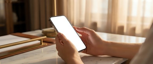 Person using smartphone with blank white screen in cozy bedroom setting with warm natural lighting and books on bedside table for mockup design.