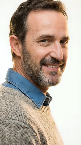 Middle-aged man with dark beard and facial hair wearing denim shirt over tan sweater, smiling at camera against white background for portrait use.