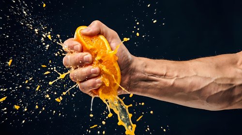 Hand squeezing orange with juice splashing against dark background. Dynamic citrus fruit action shot for beverage, health and food advertising campaigns.
