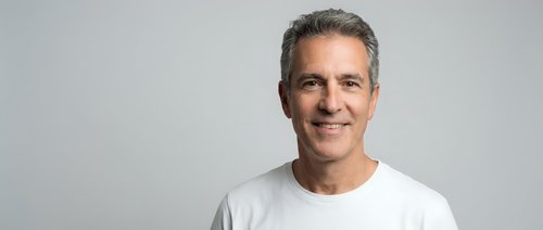 Confident mature Caucasian man with gray hair wearing white t-shirt smiling at camera on neutral background for business and lifestyle content.