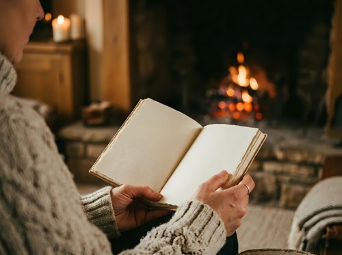 Close-up of person in cozy sweater reading open book by fireplace with warm ambient lighting and blurred flames in background.