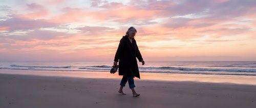 Young woman in dark winter coat walking alone on sandy beach at sunset, suitable for lifestyle, travel, and wellness editorial use.