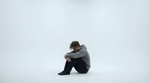 Young man sitting alone in contemplative pose against minimalist white background expressing solitude, depression, mental health struggles and emotional isolation.