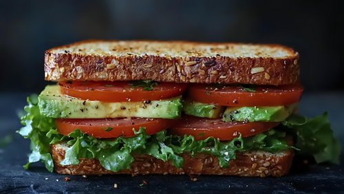 Fresh sandwich with crispy toasted bread, layered with ripe tomato slices, creamy avocado, and crisp lettuce on dark background, close-up view.