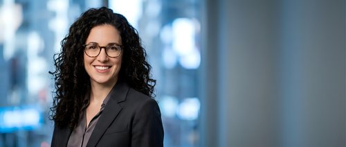 Young woman with long curly dark hair and glasses wearing professional black blazer in modern office setting with blurred city skyline background.