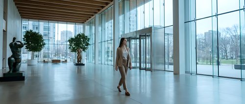 Professional businesswoman walking through modern corporate office lobby with floor-to-ceiling windows and natural lighting for business concepts.