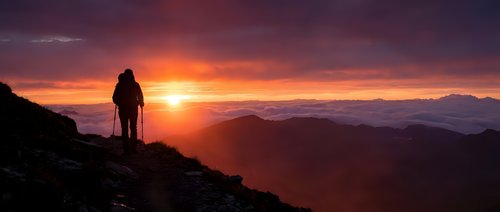 Silhouetted hiker standing on mountain ridge at golden sunrise, overlooking vast valley landscape with dramatic orange and purple sky, adventure and exploration concept.