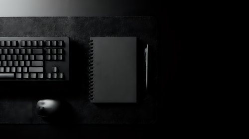Dark minimalist workspace with black keyboard, notebook, pen and mouse on textured surface for modern office branding and professional design projects.