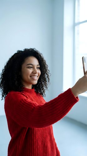 Happy young African American woman in red sweater taking selfie with smartphone near window in bright modern interior for social media content.
