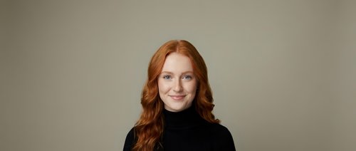 Professional redhead woman in black turtleneck sweater smiling confidently against neutral background for business and corporate marketing materials.