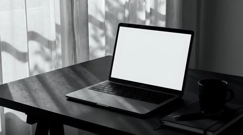 Laptop with blank white screen on dark wooden desk near window, black and white moody home office workspace with coffee mug.