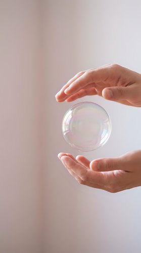 Hands holding transparent glass sphere with iridescent reflections against neutral background, symbolizing care, protection, fragility, and sustainability concepts.