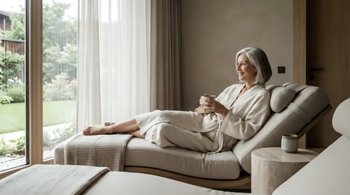 Senior woman with gray hair, relaxing on a beige upholstered lounge chair with cushions, holding a cup of tea or coffee, wearing casual cream-colored clothing, in a modern living room with large windows overlooking a garden.