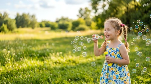 Young girl with blonde hair in braids, wearing a colorful floral dress, blowing bubbles outdoors in a sunny meadow surrounded by green grass and trees.