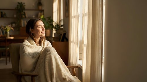 Young woman relaxing in cozy knit blanket by sunny window at home enjoying peaceful morning light and warmth in comfortable living space.