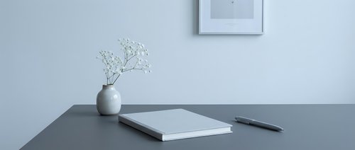 Minimalist workspace with notebook, pen, and white flower vase on gray desk, framed art on wall, clean modern home office aesthetic.