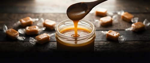 Homemade salted caramel sauce dripping from wooden spoon into glass jar surrounded by golden caramel candies on dark rustic surface for dessert recipes.