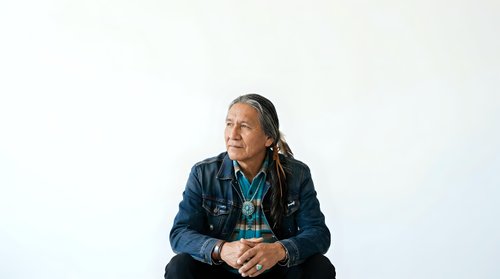 Middle-aged man with long dark hair and Native American ethnicity wearing layered denim and blue shirt, sitting with hands clasped, isolated on white background for portrait and character design use.