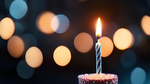 Single lit birthday candle on cupcake with sprinkles against dark background with soft glowing bokeh lights creating festive celebration atmosphere.