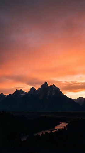 Dramatic mountain peaks silhouetted against vibrant orange and purple sunset sky with winding river valley below creating stunning landscape scenery.