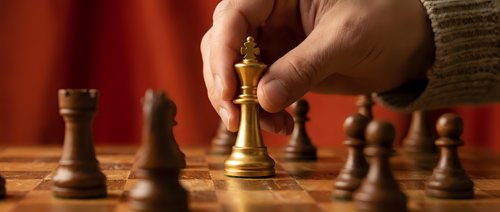 Hand moving golden chess king piece on wooden board during strategic game with warm lighting and red background creating focused atmosphere.