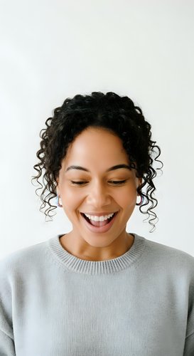 Young woman with curly black hair and medium brown skin tone wearing light gray sweater, smiling with eyes closed against white background for portrait and lifestyle use.