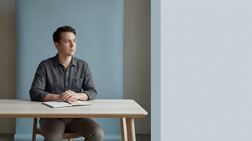 Young professional man in casual shirt sitting at modern wooden desk in minimalist office space with soft natural lighting for business presentations.