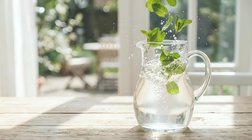 Fresh mint leaves splashing into clear water pitcher on wooden table by sunny window creating refreshing summer drink with natural lighting and bokeh background.