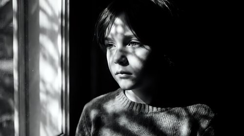 Young woman with dark hair looking pensively through window with dramatic shadows and light patterns creating moody black and white portrait atmosphere.
