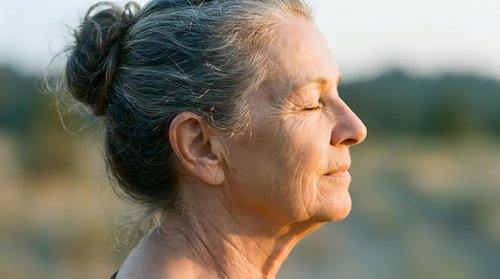 Senior woman with gray hair in bun, peaceful expression, outdoor natural light, contemplative portrait for wellness and aging themes.