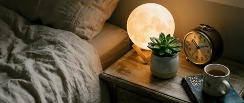 Cozy bedroom nightstand with glowing moon lamp, succulent plant, alarm clock and coffee mug creating warm ambient lighting for relaxation and sleep.