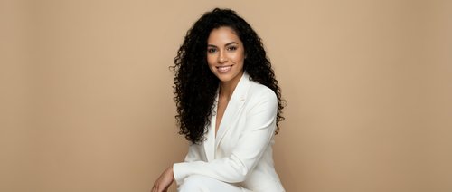 Professional Hispanic businesswoman in white blazer suit smiling confidently against neutral beige background for corporate marketing and business presentations.
