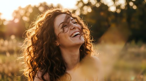 Young woman with curly hair laughing joyfully in golden sunset light outdoors. Natural happiness and positive emotions in warm evening glow.