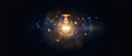 Glowing light bulb with warm filament against dark background with blue and golden bokeh lights for innovation and creativity concepts.