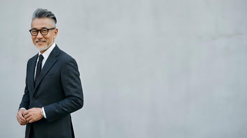 Mature male professional with gray hair and glasses wearing dark business suit and tie, standing confidently against neutral background for corporate portraits and business communications.