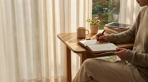 Woman in cozy beige sweater writing in journal at wooden desk by window with warm morning light, coffee cup and plant creating peaceful workspace atmosphere.